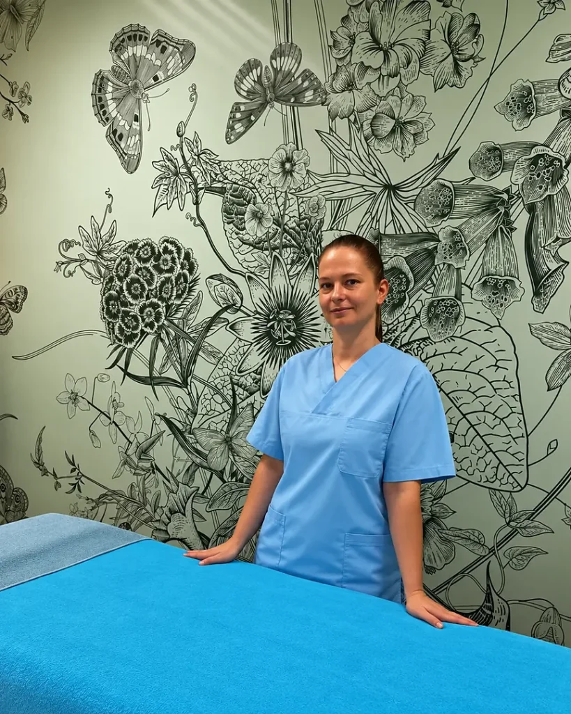 Inna steht neben der vorbereiteten Liege im Faircare-Studio und freut sich auf Ihren Besuch.
