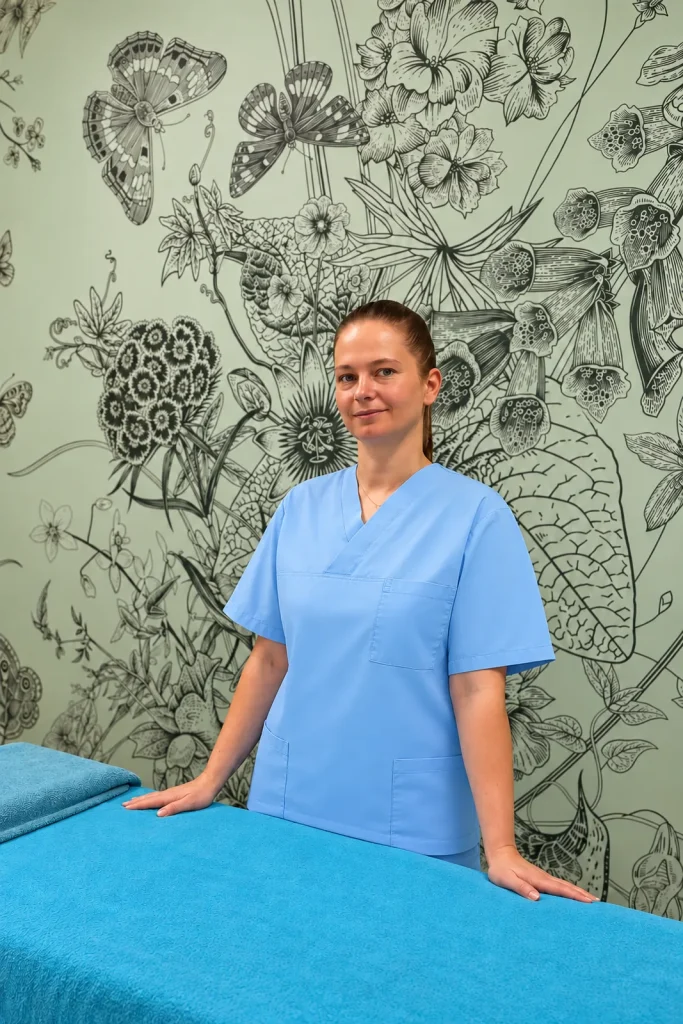 Inna in a light blue uniform stands at the turquoise treatment bed in front of the botanical wallpaper.